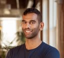 A smiling man with dark skin and dark hair wearing a navy t-shirt.